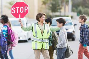School crossing guard (Hispanic mature woman, 50s) helping children walk across street. Focus on woman.
