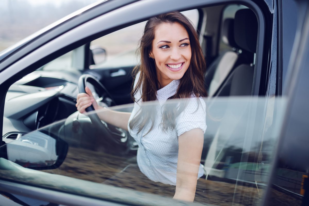 Portrait of gorgeous brunette in shirt entering her car. With one hand woman is holding steering wheel and with other shutting door.