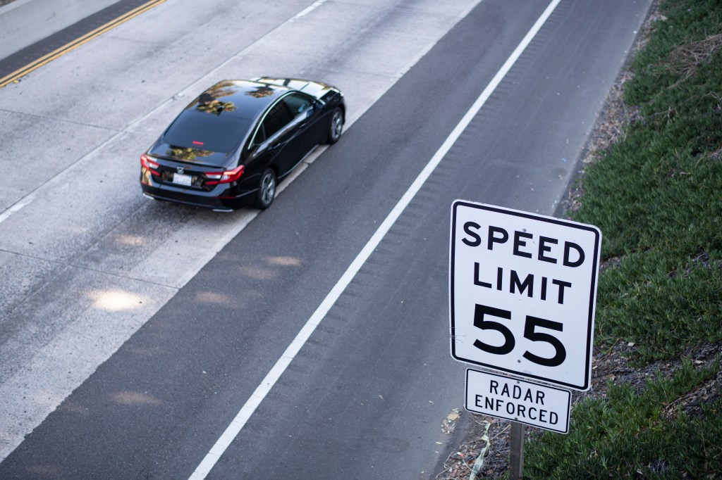 Black sedan driving on the freeway with 55 mph speed limit sign.