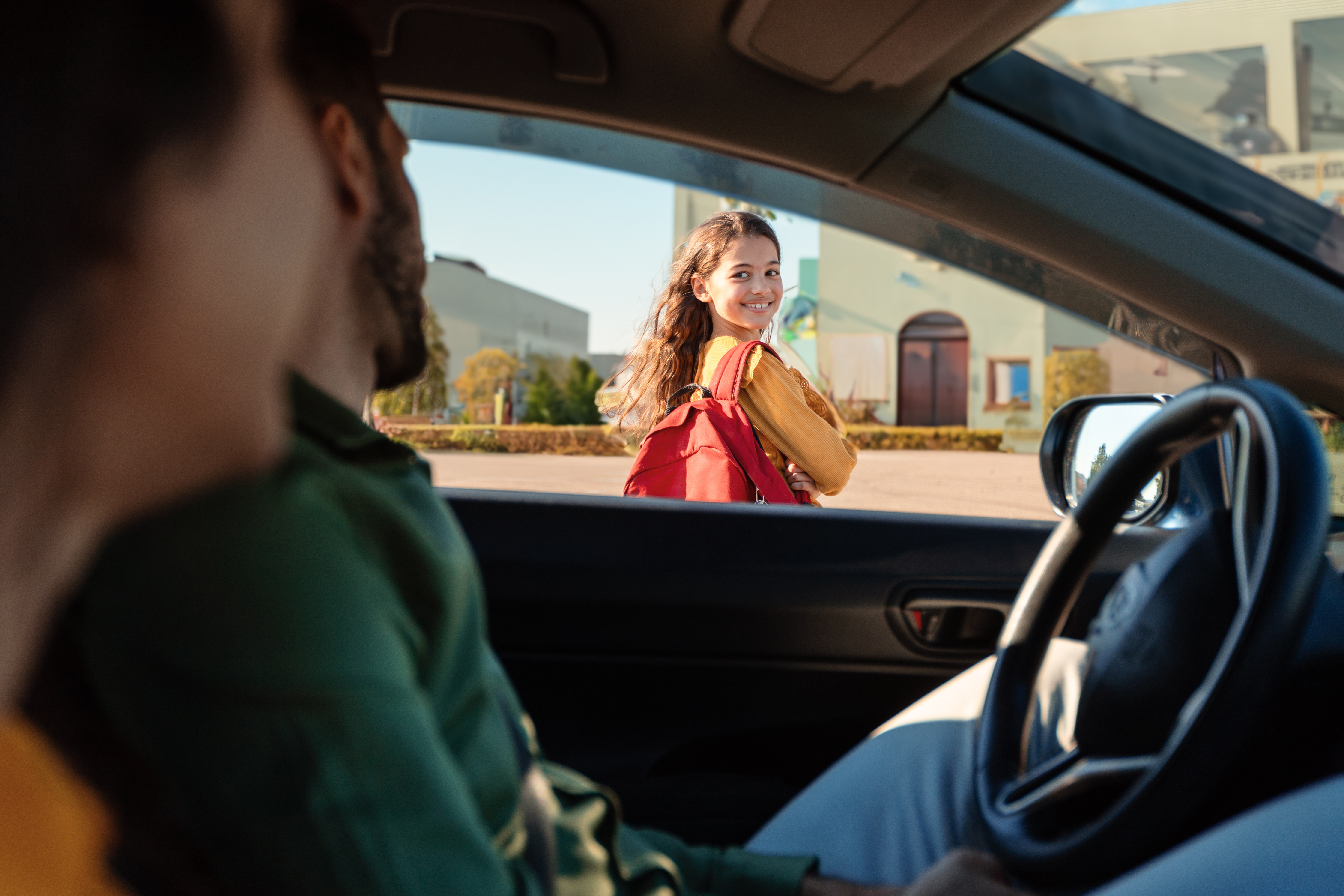 Mother and father dropping off daughter in front of school, schoolgirl turning back to parents sitting in car and smiling, view from inside auto