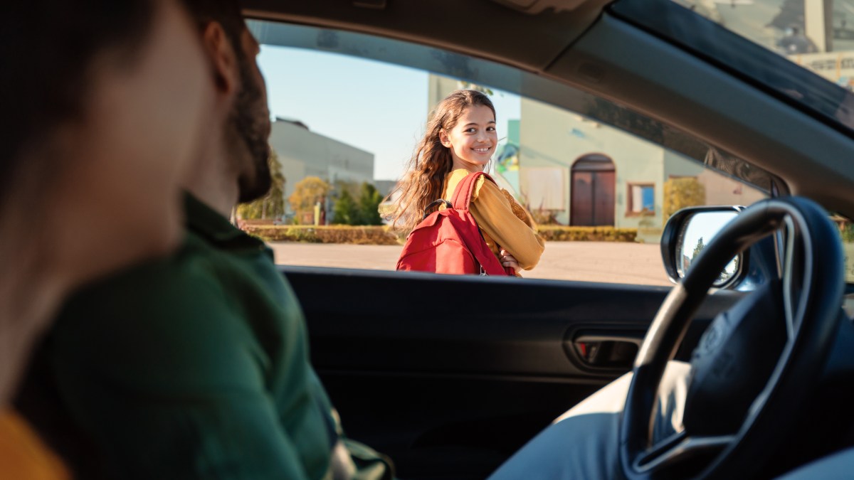 Mother and father dropping off daughter in front of school, schoolgirl turning back to parents sitting in car and smiling, view from inside auto